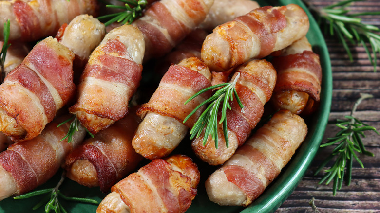 Bacon-wrapped sausages sitting on a plate with rosemary twigs sprinkled around.