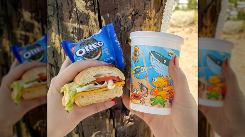 Two hands holding a small drink, sandwich, and package of Oreos