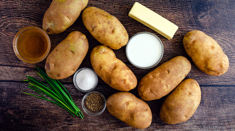 Russet potatoes, a stick of butter, chives, and containers of spices rest on a wooden surface