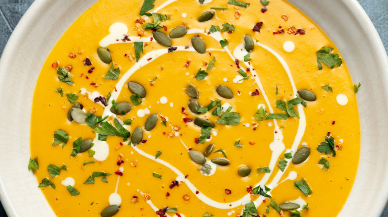 A white ceramic bowl filled with creamy butternut squash and SugarBee apple soup, topped with pepitas, red pepper flakes, parsley, and a drizzle of cream