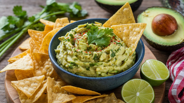 ovehead shot of bowl holding guacamole near a few chips, half of an avocado, and other ingredients to make guac