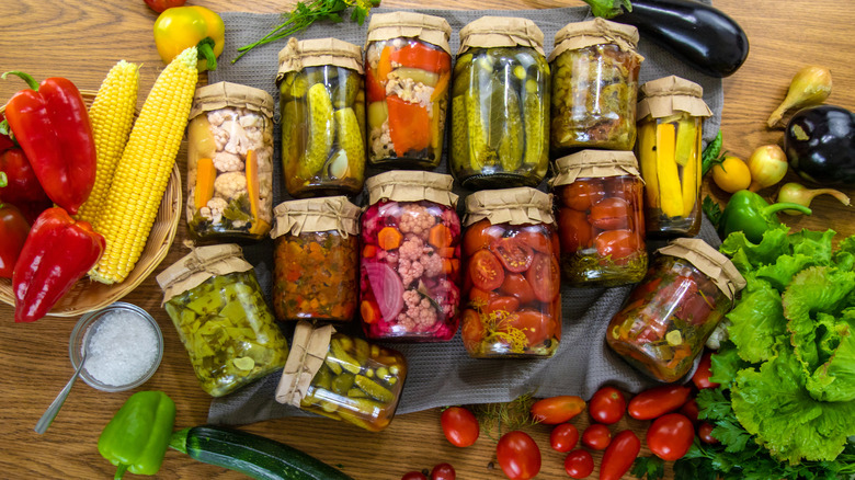 Several jars filled with canned food items lay on their side on a table. They are surrounded by vegetables.