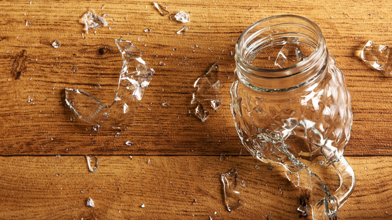 A smashed mason jar rests on a hardwood floor. Shards of glass surround it.