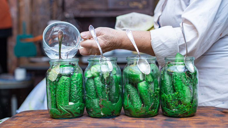 Four jars of vegetables sit on a table. Three have spoons sticking out of them. Liquid is being poured into the one on the far left.