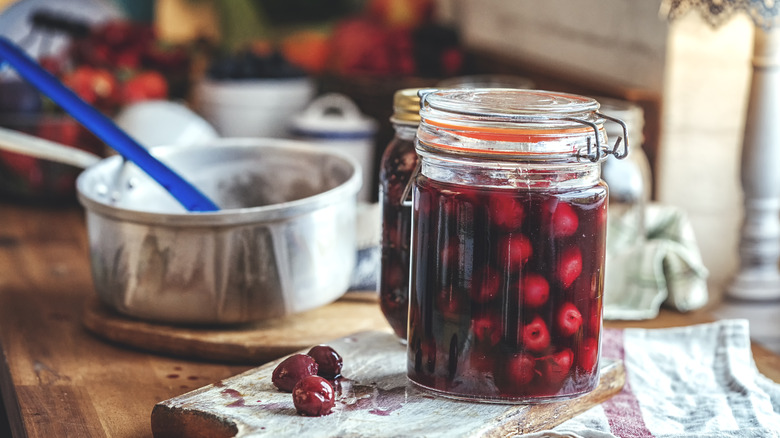 A jar filled with cherries sits on a wooden table. A bowl and various kitchen items sit in the background.