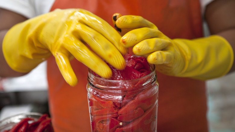 A person with yellow rubber gloves and an orange apron is placing peppers in a mason glass.