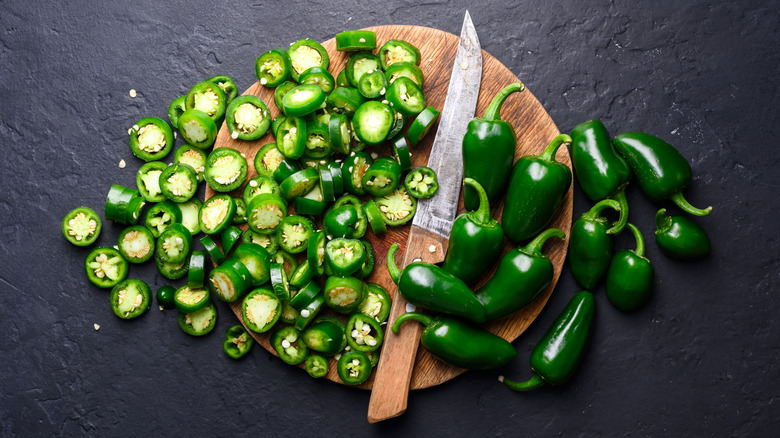 Whole and sliced green jalapeno hot peppers with knife on wooden cutting board close up.