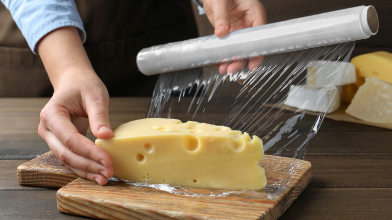 A piece of cheese on a cutting board, with hands wrapping plastic wrap around it