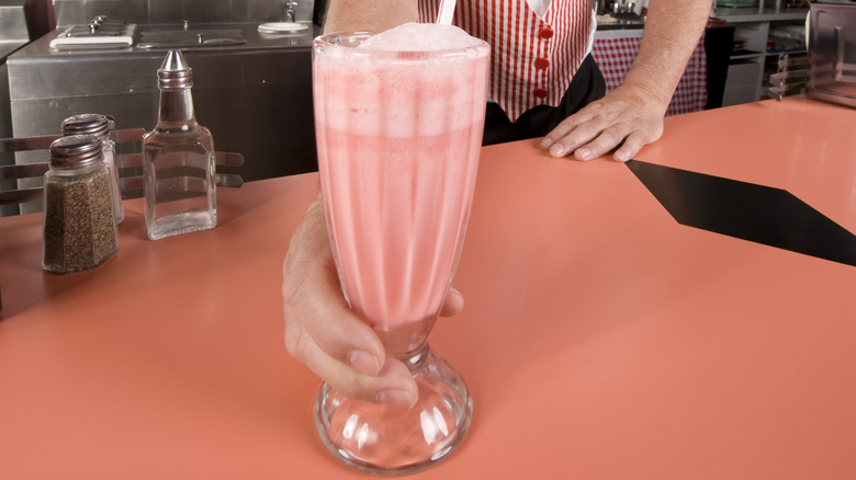 A hand extends a pink milkshake with a straw across a countertop