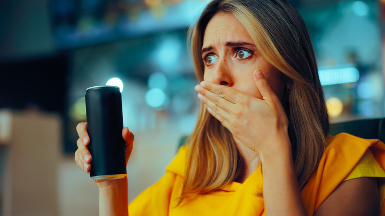 Woman holding up black aluminum can with hand over mouth and shocked face