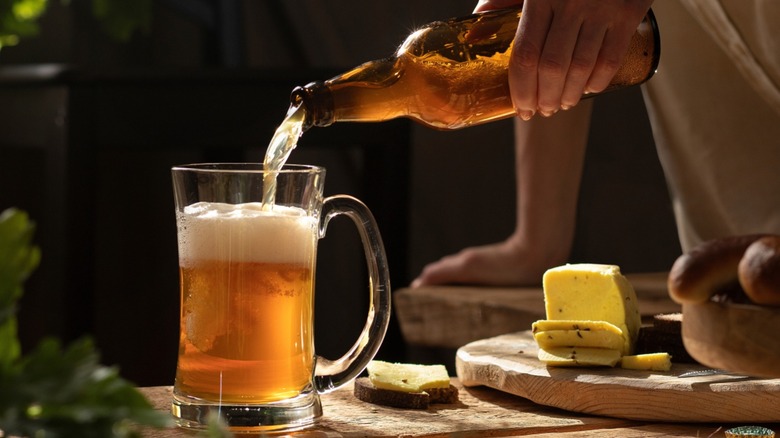 A person pouring a mug full of beer with a sliced block of cheese nearby