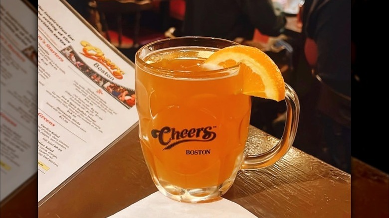A drink in a glass mug from Cheers Beacon Hill, in Boston, with an orange garnish and a menu