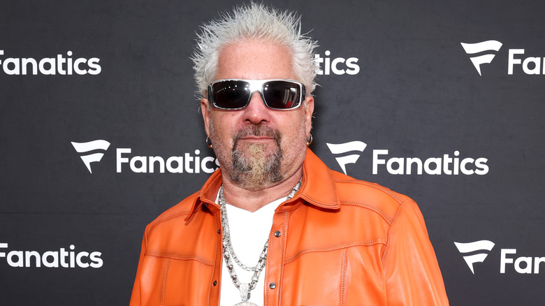 Guy Fieri wearing an orange jacket and sunglasses at a Fanatics event.