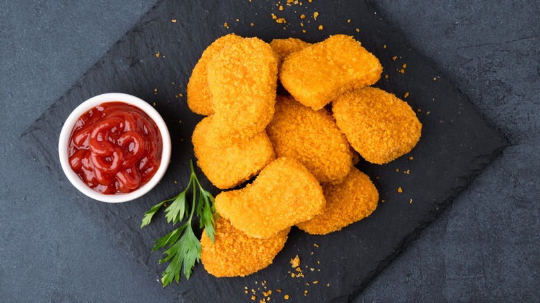 A pile of chicken nuggets lay on a black background next to a cup of ketchup