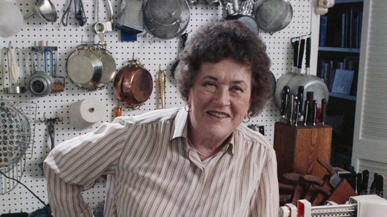 Julia Child poses for the camera in her Santa Barbara kitchen, with several utensils dangling from a pegboard wall behind her.