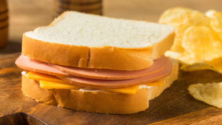 Bologna sandwich with cheese and white bread on a wooden board with chips in the background.