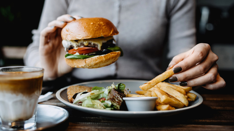 A person holding a cheeseburger in one hand and a french fry in another over a plate with a glass of coffee next to it