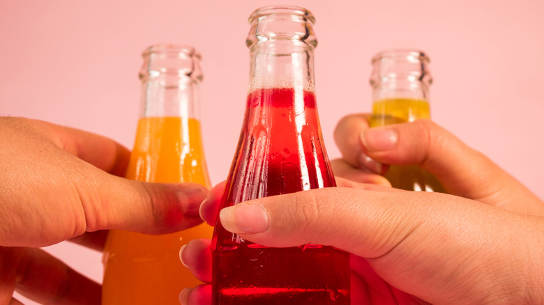 Hands clinking three soda bottles, two with orange and one with red liquid, against a pink backgrround