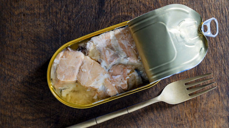 Open tin can of salmon fish fillet in its own juice and metal fork on wooden table background.