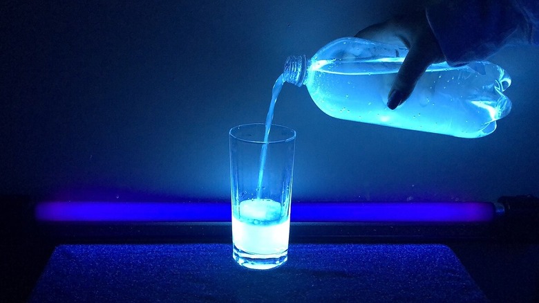 A hand pours a bottle of tonic water, which is illuminated by ultra-violet light, into a glass in a dark room