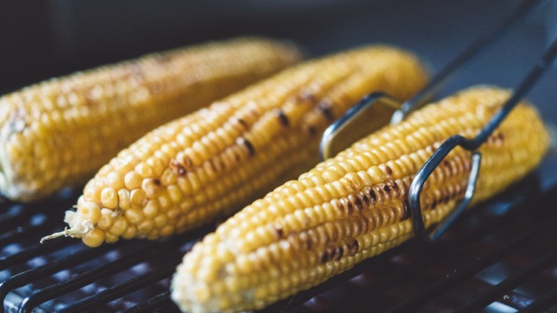 Three ears of corn being turned with pair of tongs on a grill.
