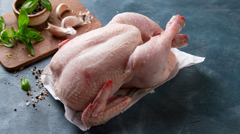 Raw whole chicken on paper with herbs on cutting board nearby