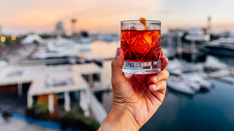 A man holding up a Negroni cocktail in front of a boat dock