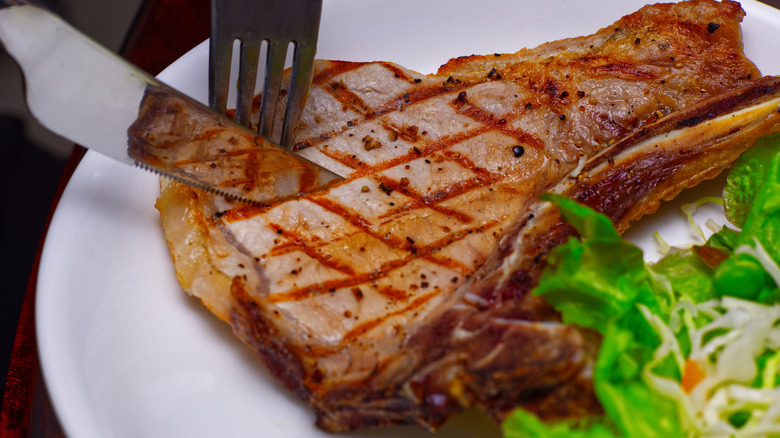 Fork and knife cutting into grilled pork chop on white plate