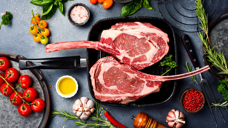 Rare tomahawk steaks in a fry pan surrounded by herbs and seasonings