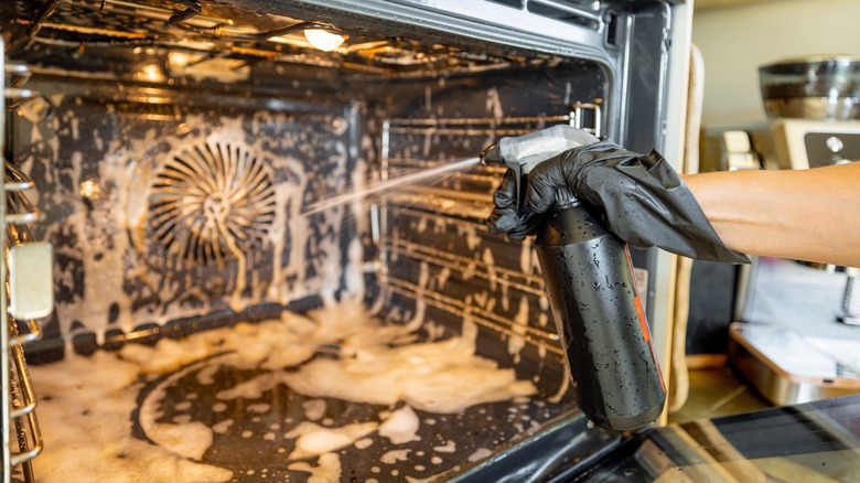 A close-up of a hand in a black glove spraying cleaning solution inside a dirty oven.