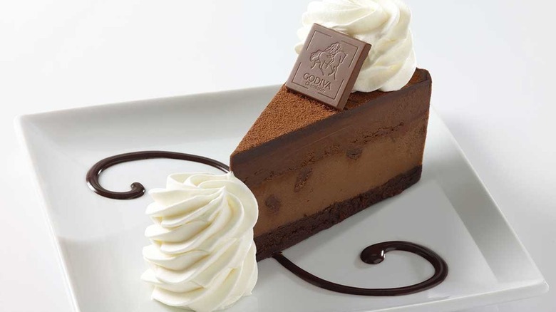 slice of Godiva Chocolate Cheesecake from The Cheesecake Factory on a square white plate decorated with a chocolate sauce drizzle and a pile of whipped cream