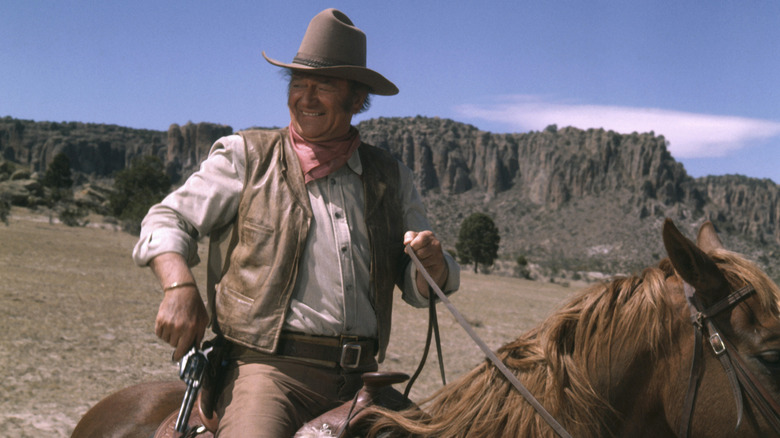 John Wane on horseback smiling while holding a revolver.