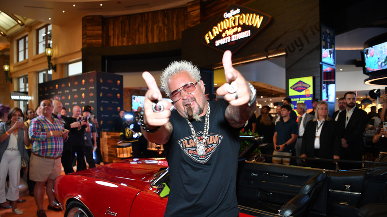 Guy Fieri poses in front of his iconic car, with the "Flavortown" sports kitchen sign in the background