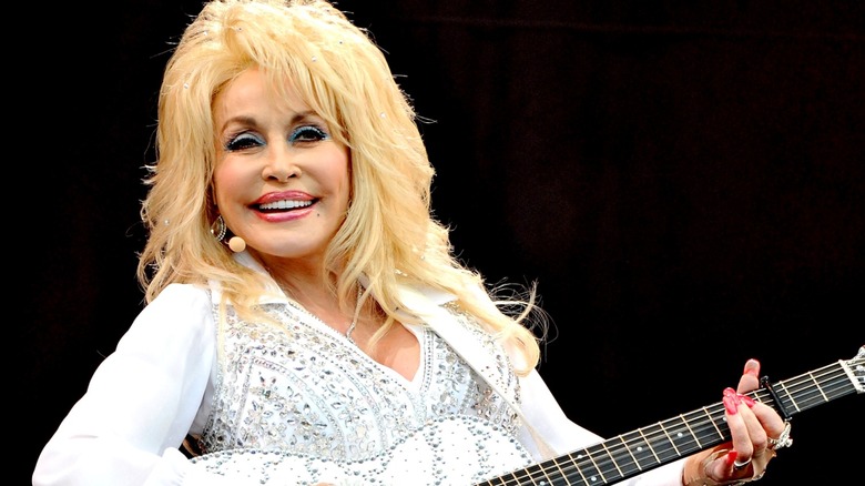 Dolly Parton smiling in white rhinestone-covered outfit playing guitar