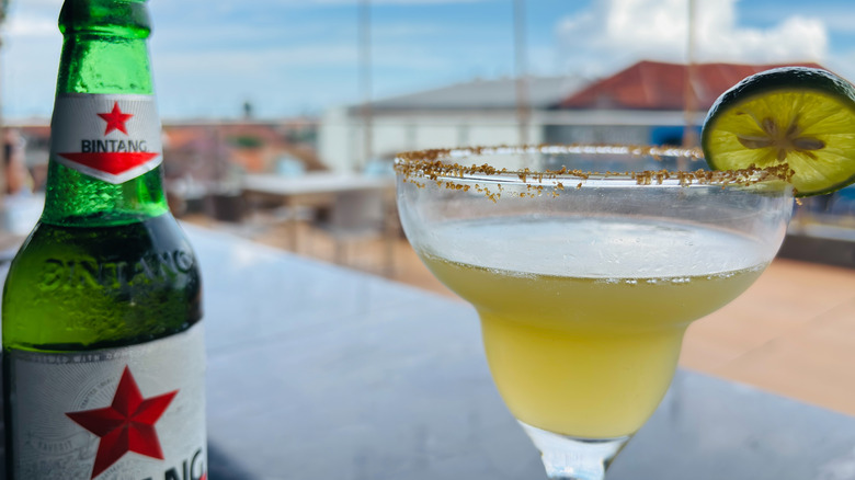 A green bottle of foreign beer next to a greenish-yellow margarita on a table outside