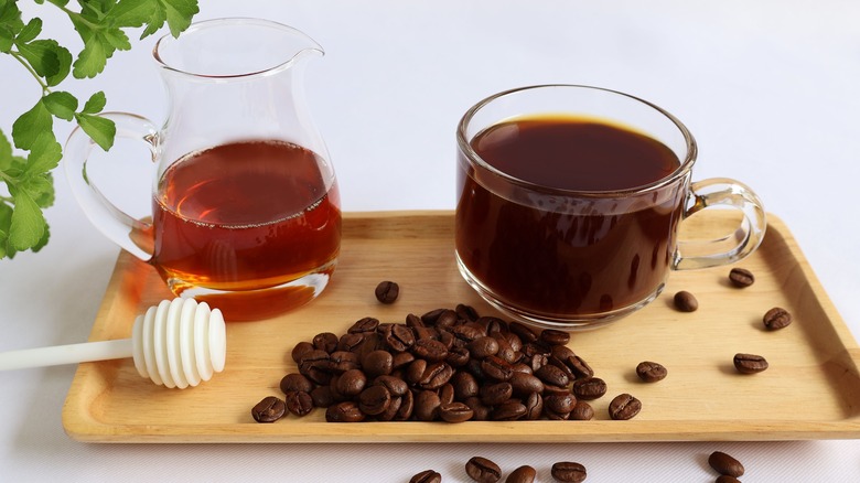 Honey container and cup of coffee on a wooden tray, with coffee beans spread around.