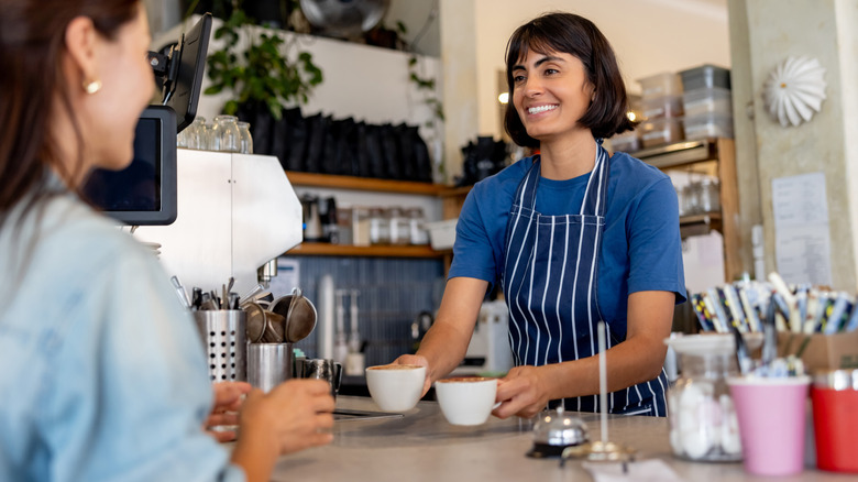 A barista hands two ceramic mugs across a bar to a customer at a coffee shop