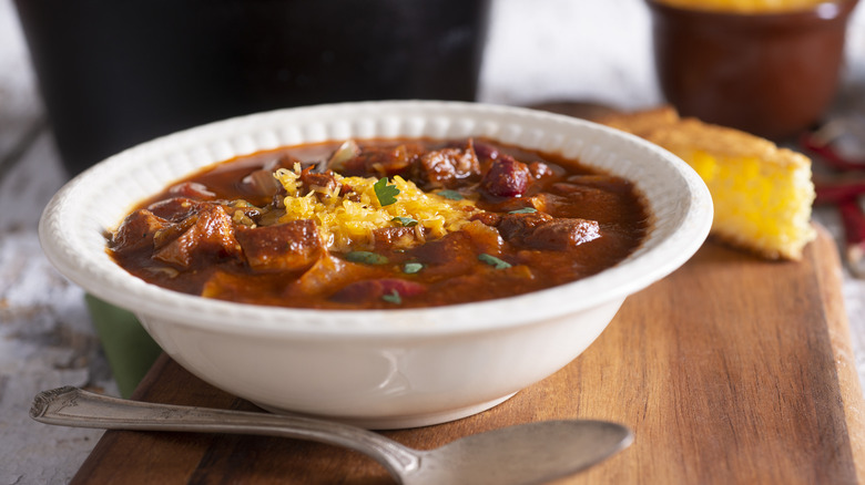 Bowl of chili with cheese, spoon, cornbread slice on board