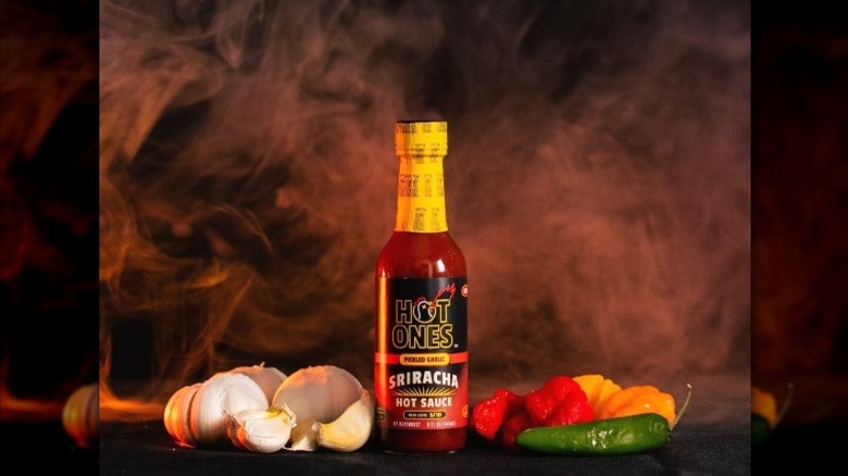 A bottle of Hot Ones Pickled Garlic Sriracha Hot Sauce with dramatic smoke and ingredients.
