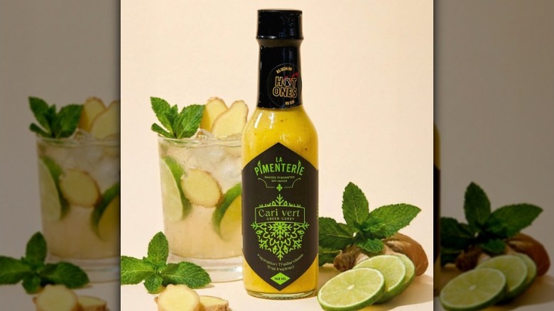 A bottle of La Pimenterie Green Curry Hot Sauce next to a cocktail and sliced limes.
