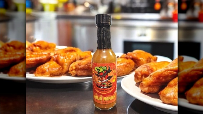 A bottle of The Pepper Ninja Ninja Napalm Hot Sauce in front of plates of wings.