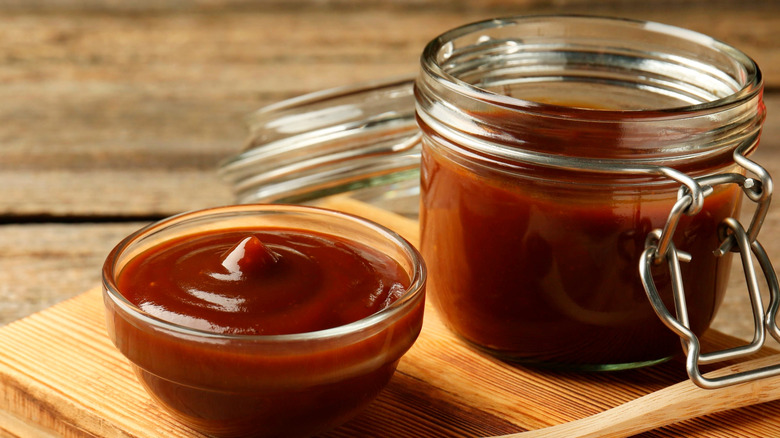 A glass jar of BBQ sauce next to a small glass bowl of BBQ sauce.