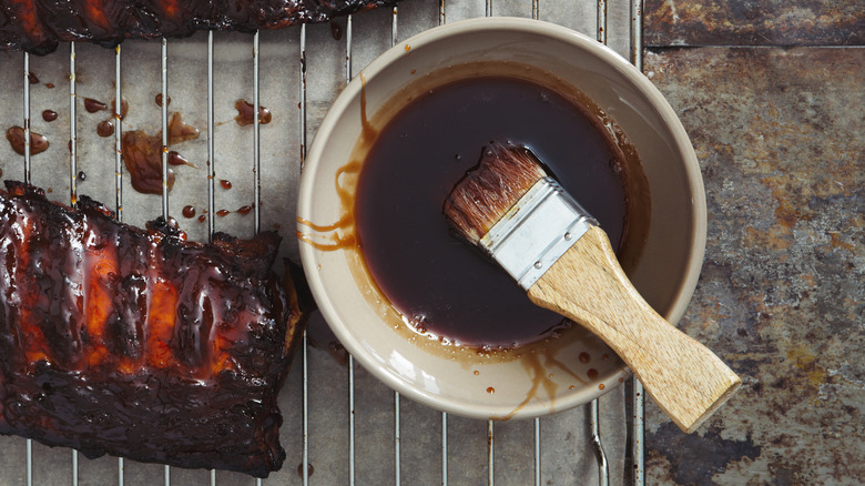 A top-down view of a white bowl full of homemade BBQ sauce with a brush next to some ribs