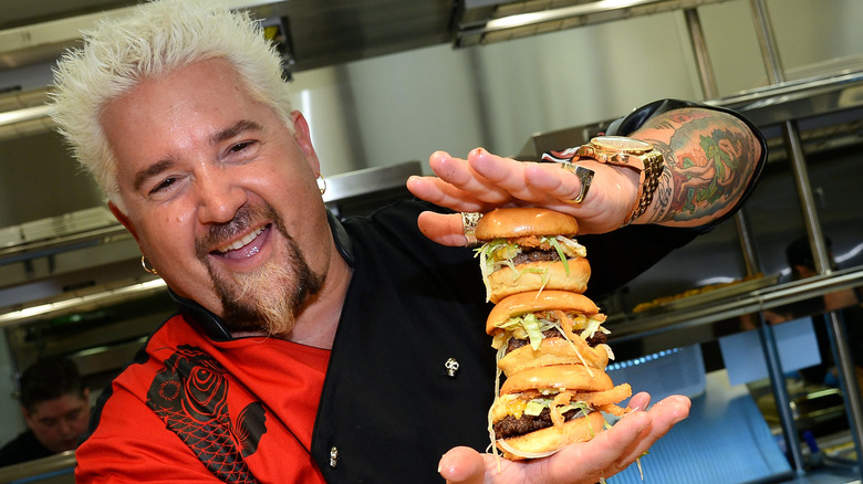 Guy Fieri holding three burgers together while smiling with his mouth open in a kitchen.