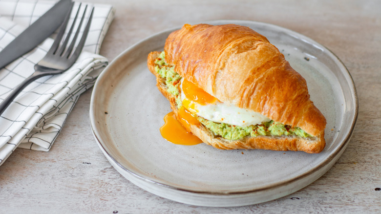 Croissant breakfast sandwich filled with smashed avocado and a runny egg served on a grey plate