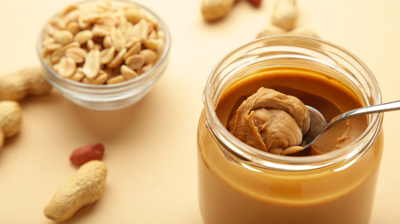 An open jar of peanut butter with a spoon and a bowl of peanuts
