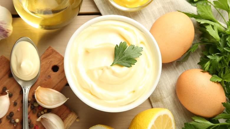 Bowl of homemade mayonnaise surrounded by eggs, garlic, parsley, lemon, and oil