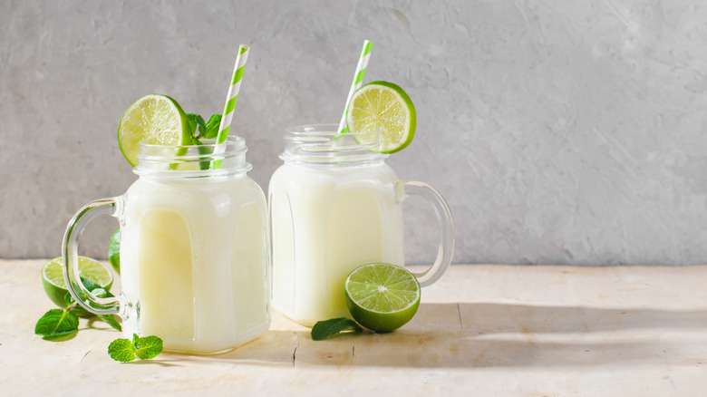 Creamy limeade in square mugs garnished with lime slices and green and white straws with two cut limes and mint springs around them on a cream colored surface