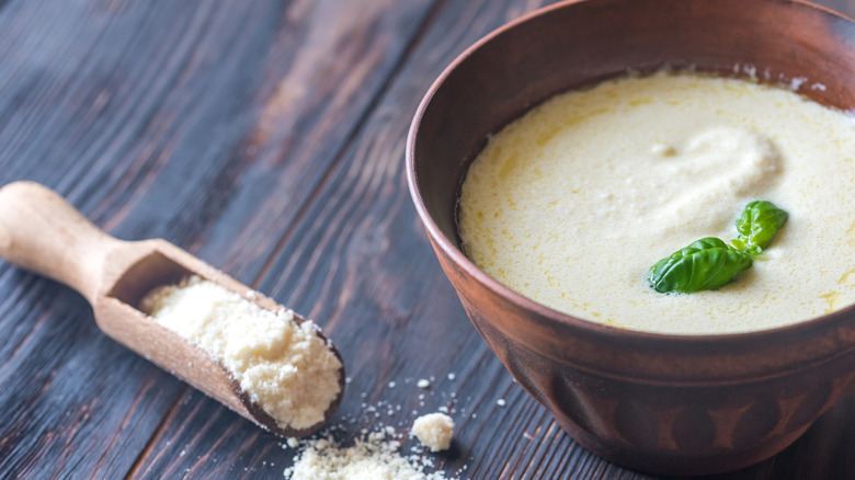 A bowl of alfredo sauce topped with a basil leaf, next to a wooden spoonful of parmesan cheese
