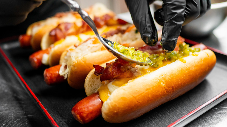 A chef adds toppings to a row of bacon and cheese hot dogs on a platter